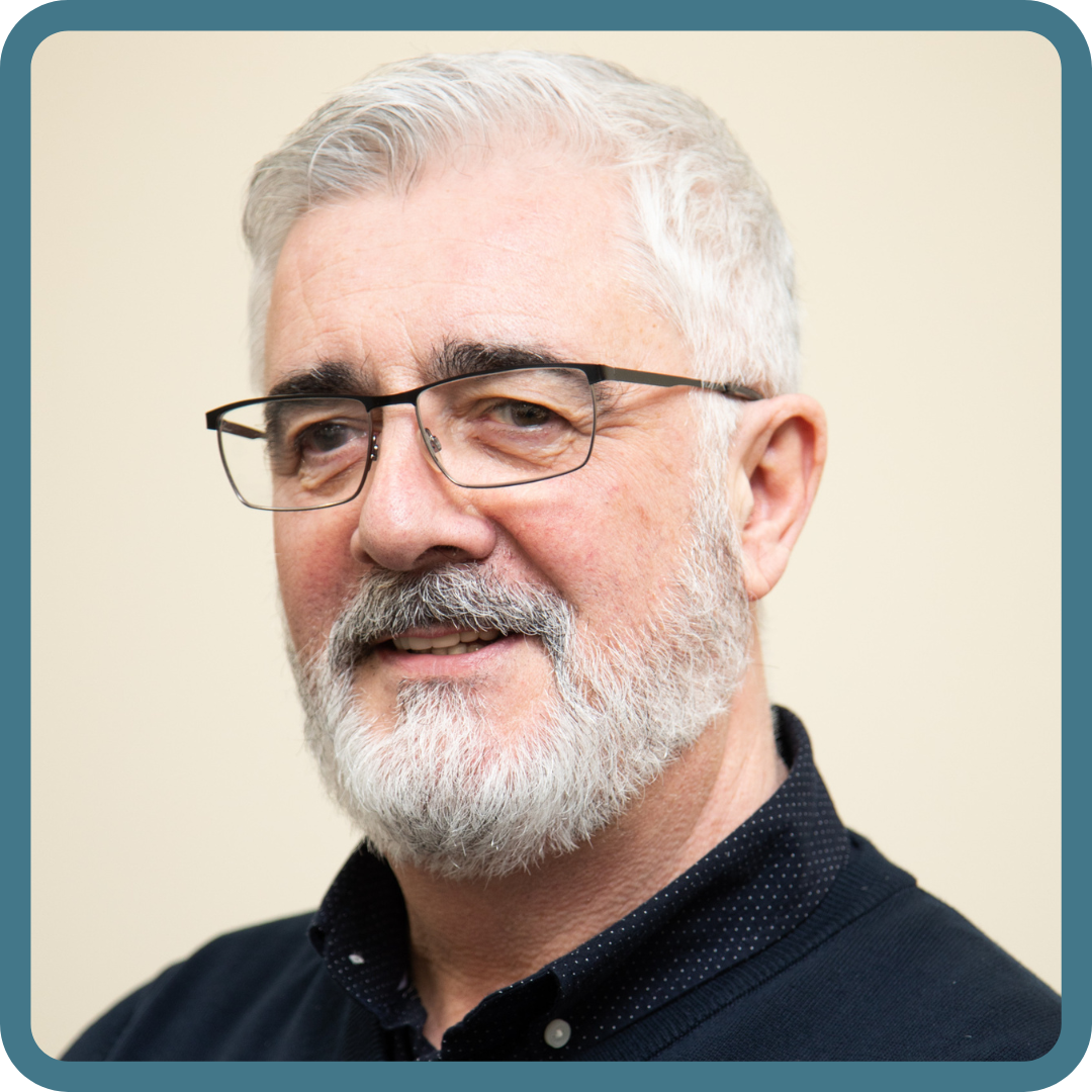 Man with glasses and grey hair smiles at camera. Wearing blue shirt. 