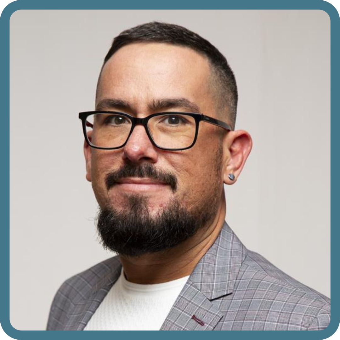Man with black glasses and black hair smiles at camera. Wearing grey suit jacket. 