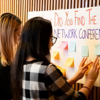 Two women stand at a board reading post it notes. 
