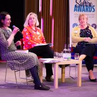 Three women sit on stage with microphone speaking to the crowd.