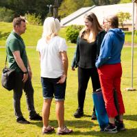 Four people stand talking in grass area. 