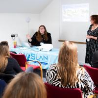Two women stand in front of a screen presenting to a group of people. 