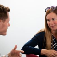 Two people are seen mid-discussion sitting down. 