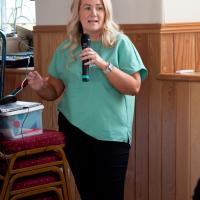 Woman with blonde hair and green t-shirt speaks to the crowd through a microphone. 