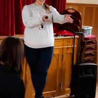 Woman with brown hair and grey jumper presents to the room. 