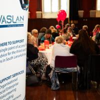 A white VASLan banner is in focus as the rest of a busy hall is out of focus. 