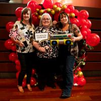Three women smile in front of red balloon display. They are holding 80s props.