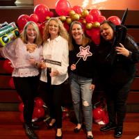 Four women smile in front of red balloon display. They are holding 80s props.