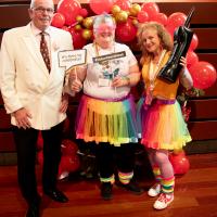 Three people smile in front of red balloon display. They are holding 80s props and dressed in 80s clothing. 