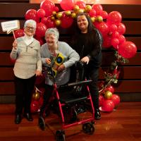 Three women smile in front of red balloon display. They are holding 80s props.