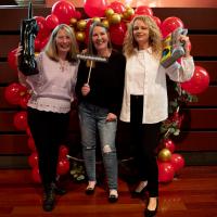 Three women smile in front of red balloon display. They are holding 80s props.