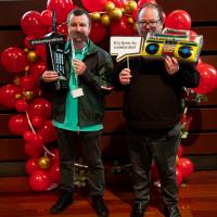 Two men smile in front of red balloon display. They are holding 80s props.