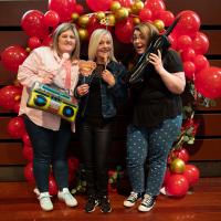 Three women smile in front of red balloon display. They are holding 80s props.